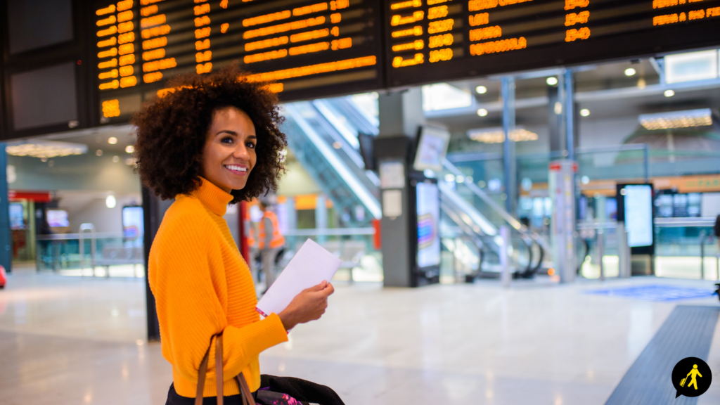 A woman wearing an orange jumper stands in front of an airport departure board with a boarding pass in her hand and a bag over her arm.