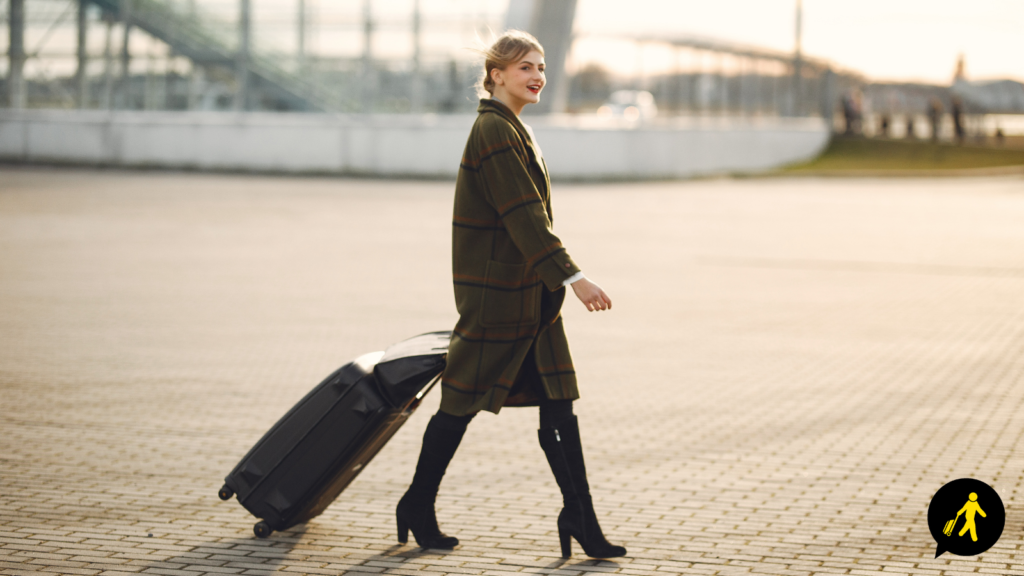 A woman in a long cat walks outside of an airport, pulling luggage behind her.