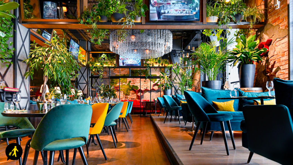 Restaurant with colourful, velvet seating and wood floors. There is a chandelier in the centre of the ceiling, exposed brick walls and plants everywhere.